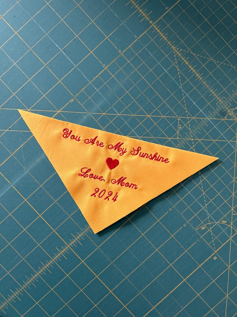 Op de afbeelding: Een gele driehoekige stof met rode geborduurde tekst die "You are my sunshine, Love, Mom 2024" en een rood hart zegt.