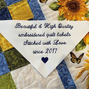 Op de afbeelding: Witte driehoekige stoffen label met blauwe geborduurde tekst die "Beautiful & High Quality embroidered quilt labels Stitched with Love since 2017" luidt. Een blauw hart is geborduurd onder de tekst. Het label bevindt zich op een witte achtergrond met een patchwork-quiltontwerp.