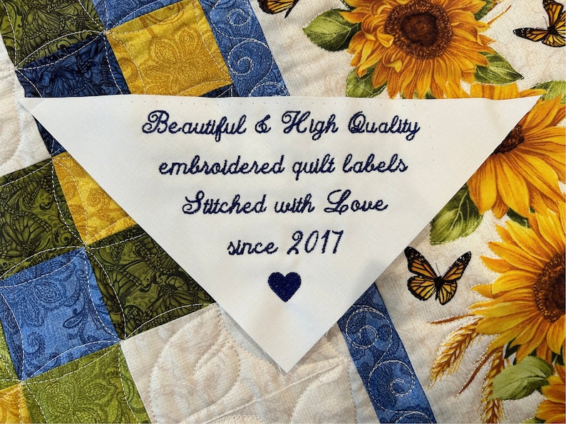 Op de afbeelding: Witte driehoekige stoffen label met blauwe geborduurde tekst die "Beautiful & High Quality embroidered quilt labels Stitched with Love since 2017" luidt. Een blauw hart is geborduurd onder de tekst. Het label bevindt zich op een witte achtergrond met een patchwork-quiltontwerp.