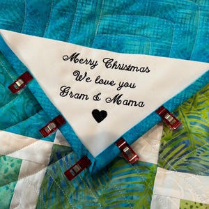 Op de afbeelding: Een wit vierkant van stof met zwarte borduurwerk dat "Merry Christmas We love you Gram & Mama" zegt met een klein zwart hart in het midden. Het vierkant van stof is vastgemaakt aan een blauwe en groene gepatroneerde quilt.