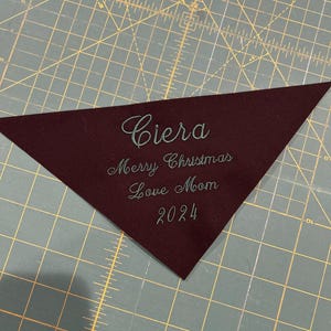 Op de afbeelding: Een bordeauxrode driehoek van stof met de naam "Ciera" geborduurd in tealgroen garen. De tekst "Merry Christmas Love Mom 2024" is ook geborduurd in tealgroen garen.