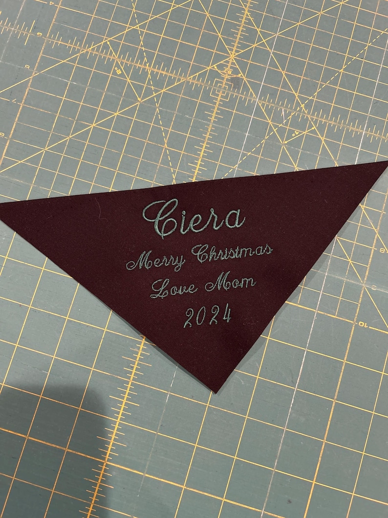 Op de afbeelding: Een bordeauxrode driehoek van stof met de naam "Ciera" geborduurd in tealgroen garen. De tekst "Merry Christmas Love Mom 2024" is ook geborduurd in tealgroen garen.