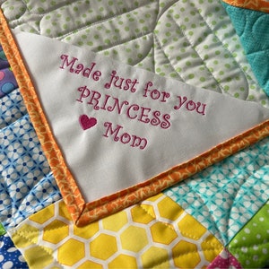 May include: A white fabric square with orange trim, embroidered with the text "Made just for you Princess Mom" in pink thread. The fabric square is attached to a colorful patchwork quilt.