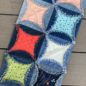 May include: A denim quilt runner with a patchwork design featuring various fabrics with floral patterns. The quilt runner is made with frayed denim edges and has a rustic, handmade look.