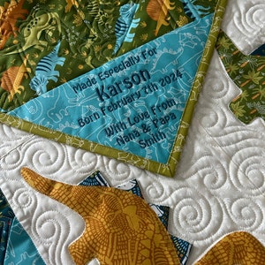 May include: A blue and green fabric label with the text "Made Especially For Karson Born February 7th 2024 With Love From Nana & Papa Smith" sewn on. The label is attached to a green and yellow dinosaur-themed quilt.