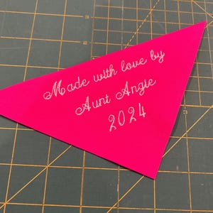 Op de afbeelding: Een roze driehoekig stoffen label met witte borduurwerk dat "Made with love by Aunt Angie 2024" zegt.