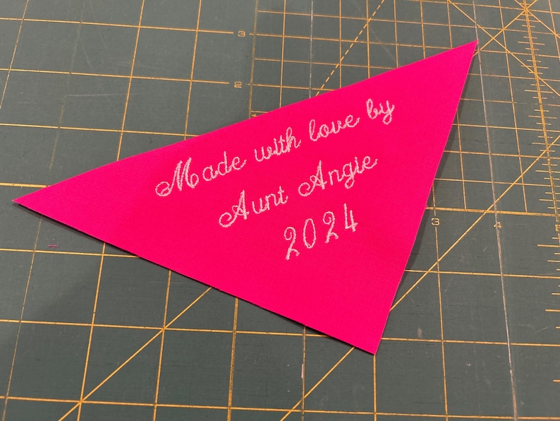 Op de afbeelding: Een roze driehoekig stoffen label met witte borduurwerk dat "Made with love by Aunt Angie 2024" zegt.
