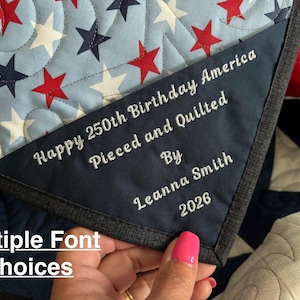 May include: A quilted fabric piece featuring a light blue background with red and navy blue stars. A dark navy corner patch is embroidered with the white text: Happy 250th Birthday America, Pieced and Quilted By, Leanna Smith, 2026. The edge has a dark trim.