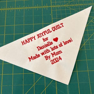 Op de afbeelding: Witte driehoekige stoffen label met rode geborduurde tekst die "Happy Joyful Quilt for Danielle Made with lots of love! By Mom 2024" en een rood hart zegt.