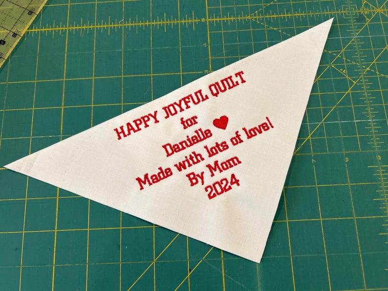 Op de afbeelding: Witte driehoekige stoffen label met rode geborduurde tekst die "Happy Joyful Quilt for Danielle Made with lots of love! By Mom 2024" en een rood hart zegt.