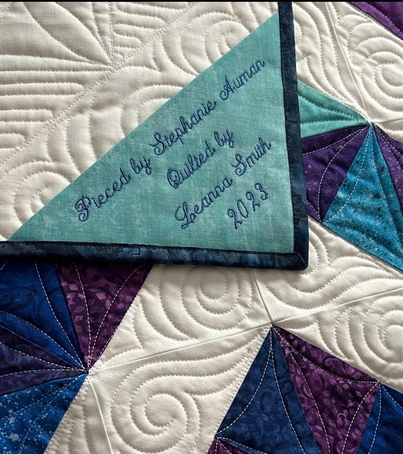 Op de afbeelding: Een wit quilt met een geometrisch patroon in blauw en paars. Het quilt is gestikt met een swirl-ontwerp. Een blauw label met witte borduurwerk staat "Pieced by Stephanie Auman, Quilted by Leanna Smith, 2023".