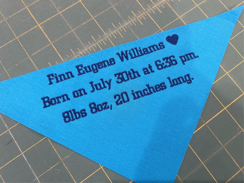 Op de afbeelding: Een lichtblauwe driehoekige stof met de tekst "Finn Eugene Williams Geboren op 30 juli om 18:36 uur. 8lbs 8oz, 20 inch lang." geborduurd in donkerblauw.