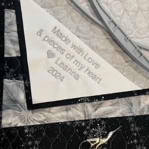May include: A close-up of a quilt with a white triangle label embroidered with the text "Made with Love & pieces of my heart Leanna 2024". The quilt has a floral pattern in shades of white and gray, with black borders and silver snowflake accents. Gold scissors are visible.