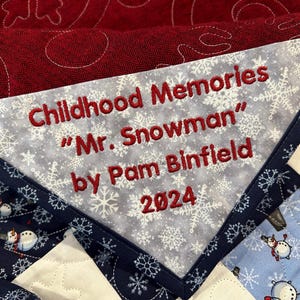 May include: A white fabric label with red embroidered text that reads "Childhood Memories "Mr. Snowman" by Pam Binfield 2024" on a snowflake patterned background. The label is attached to a blue and white fabric with a snowflake pattern.