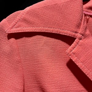 May include: Close-up of a coral-colored blazer with a textured fabric. The blazer features a notched lapel and white stitching details. The image highlights the fabric's texture and the tailored design of the garment.