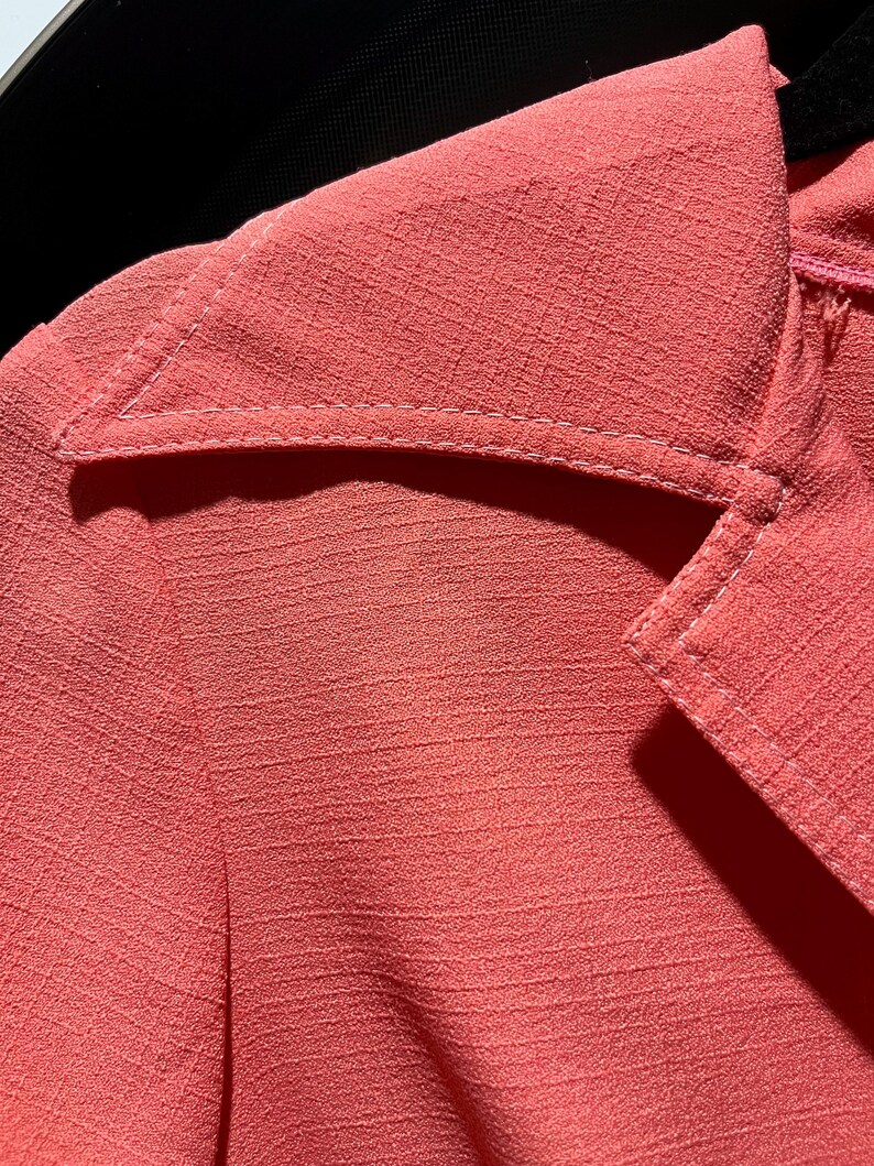 May include: Close-up of a coral-colored blazer with a textured fabric. The blazer features a notched lapel and white stitching details. The image highlights the fabric's texture and the tailored design of the garment.