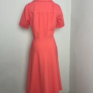 May include: A coral-colored dress with a matching short-sleeved jacket. The dress has a flared skirt and a collared neckline. The jacket has a button-up front. The outfit is displayed on a mannequin.
