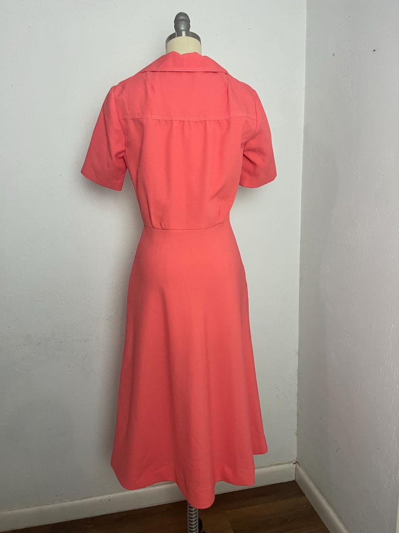 May include: A coral-colored dress with a matching short-sleeved jacket. The dress has a flared skirt and a collared neckline. The jacket has a button-up front. The outfit is displayed on a mannequin.