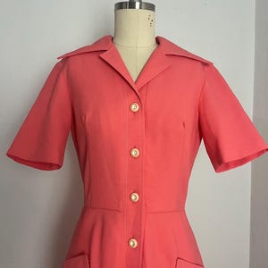 May include: Coral-colored vintage dress with short sleeves, a collared neckline, and a button-down front. The dress features two front pockets and is displayed on a mannequin. The dress is a classic style.