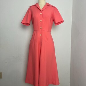 May include: A coral-colored vintage dress with short sleeves and a collared neckline. The dress features a button-down front with gold-colored buttons, two front pockets, and a midi-length skirt. The dress is displayed on a mannequin.
