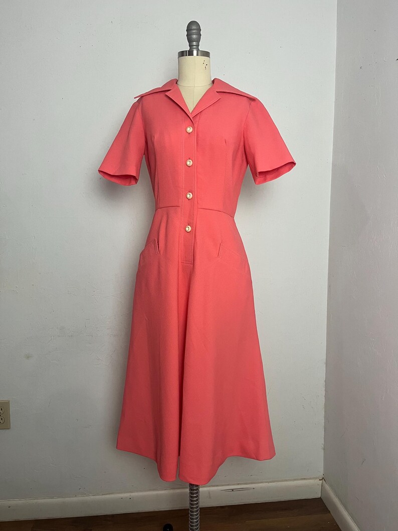May include: A coral-colored vintage dress with short sleeves and a collared neckline. The dress features a button-down front with gold-colored buttons, two front pockets, and a midi-length skirt. The dress is displayed on a mannequin.