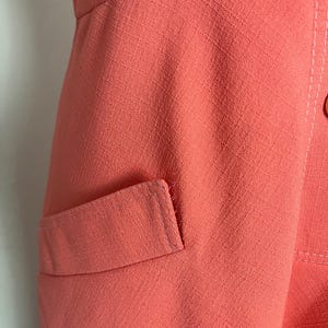 May include: Close-up of a coral-colored garment with a textured fabric. The image shows a pocket detail and stitching. The garment appears to be a dress or a top.