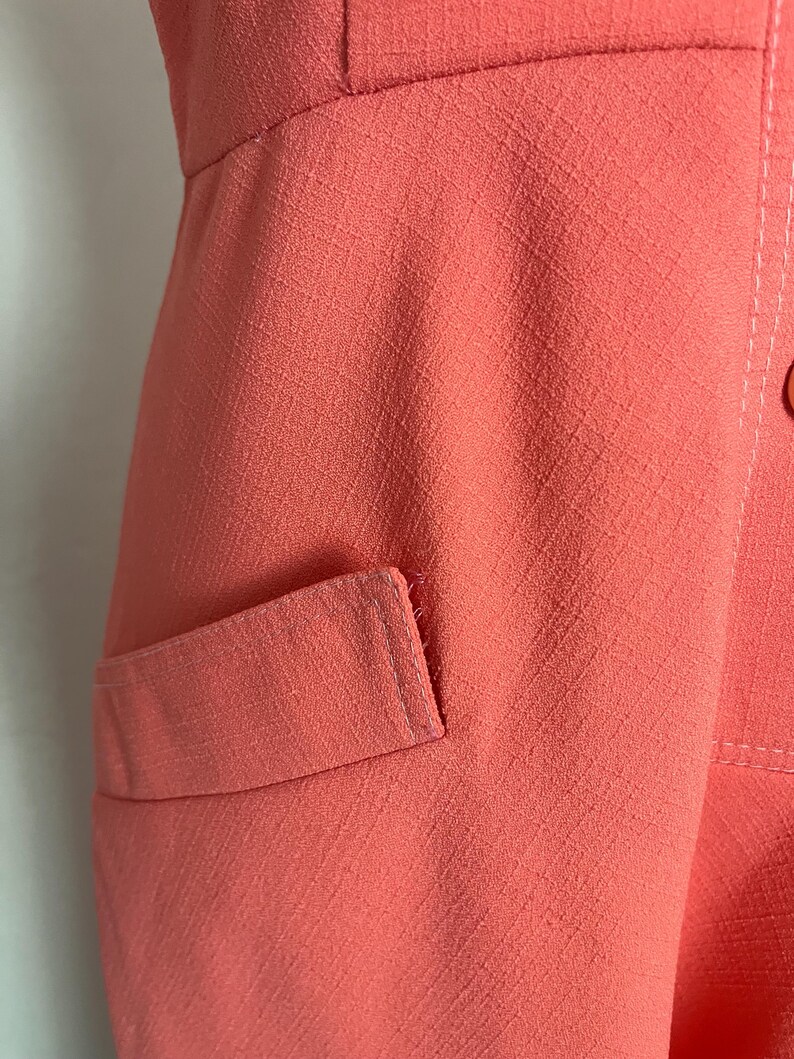 May include: Close-up of a coral-colored garment with a textured fabric. The image shows a pocket detail and stitching. The garment appears to be a dress or a top.