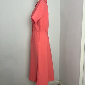 May include: A coral-colored dress with short sleeves and a collared neckline. The dress features a fitted bodice and a flared skirt, with a button-down front and pockets. The dress is displayed on a mannequin.