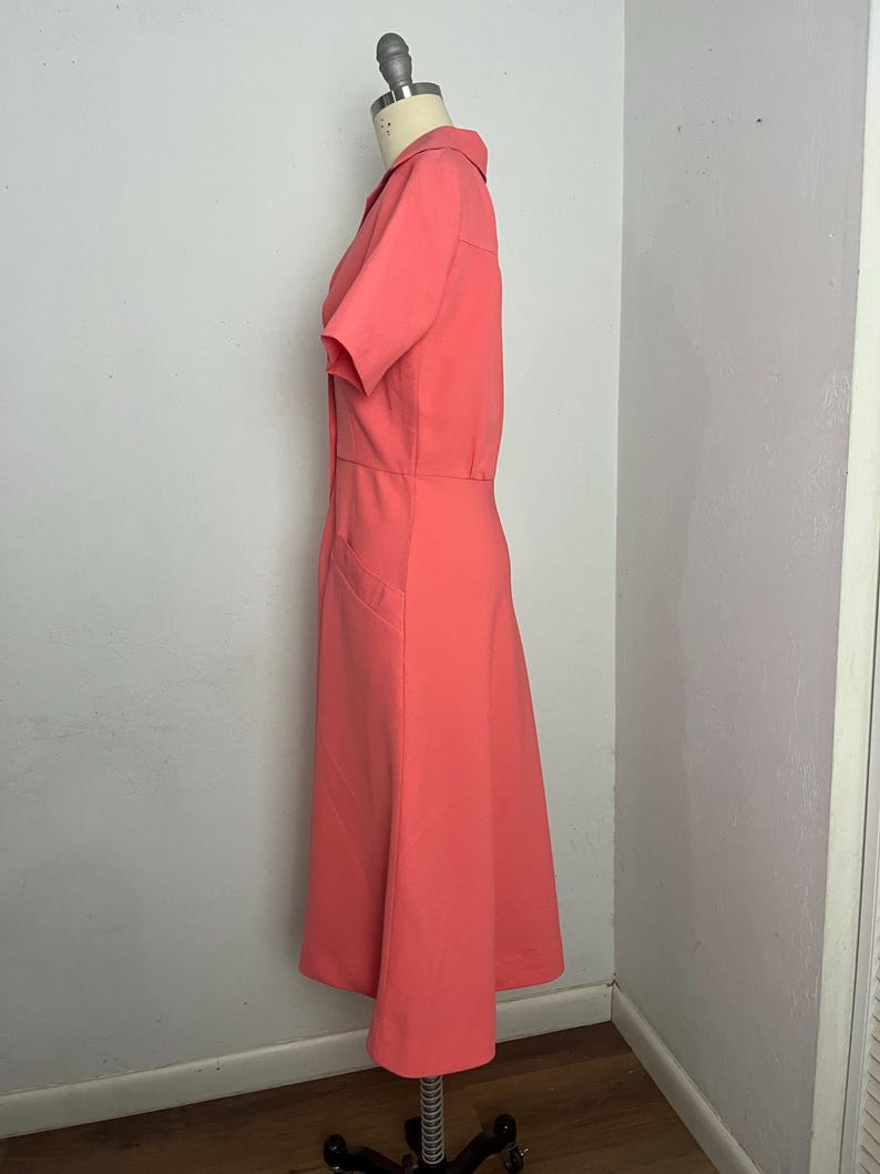 May include: A coral-colored dress with short sleeves and a collared neckline. The dress features a fitted bodice and a flared skirt, with a button-down front and pockets. The dress is displayed on a mannequin.