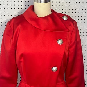May include: A vibrant red coat featuring a wide collar and three large, round, off-white buttons with silver-toned, crystal-encrusted borders. The coat has long sleeves and a structured, pleated waistline, displayed on a mannequin torso.