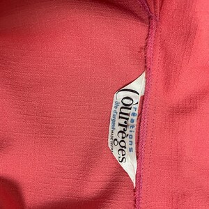 May include: Close-up of a textured coral-colored fabric with a white tag. The tag features the brand name "Courr&egrave;ges" in a stylized font, along with additional text. The fabric has a subtle, grid-like pattern.