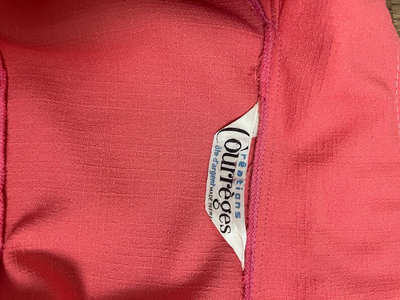 May include: Close-up of a textured coral-colored fabric with a white tag. The tag features the brand name "Courr&egrave;ges" in a stylized font, along with additional text. The fabric has a subtle, grid-like pattern.