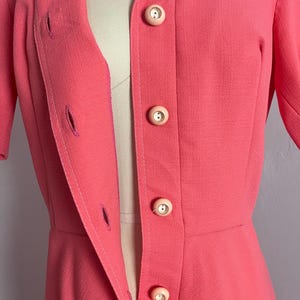 May include: A coral-colored vintage blazer with a notched collar and short sleeves. The blazer features three large, round, light pink buttons down the front and two pockets. The blazer is tailored with a fitted waist and a classic design.