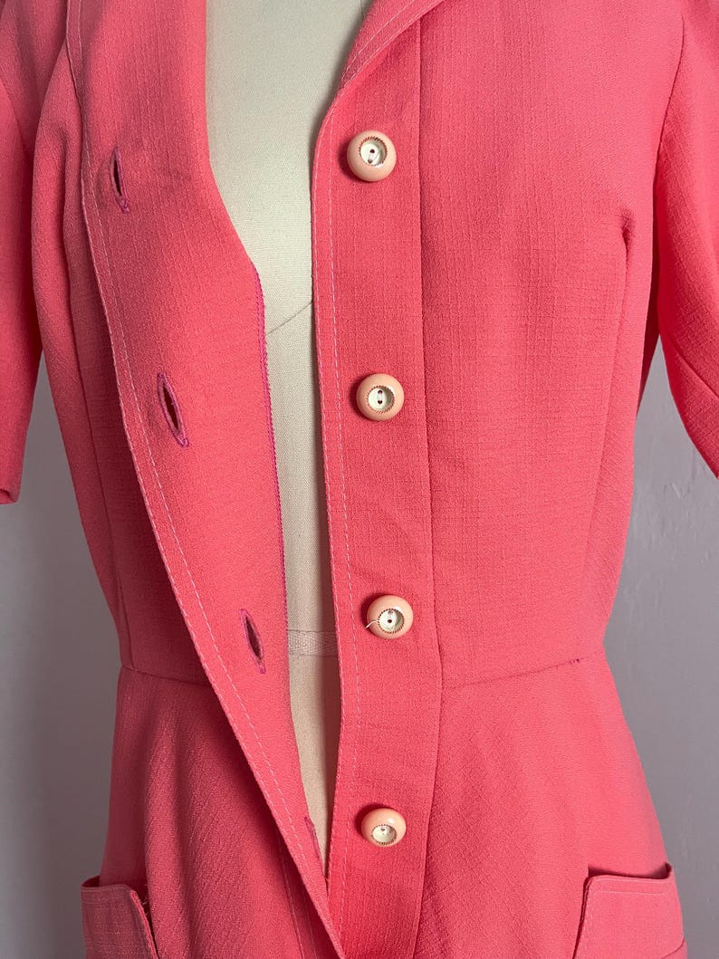 May include: A coral-colored vintage blazer with a notched collar and short sleeves. The blazer features three large, round, light pink buttons down the front and two pockets. The blazer is tailored with a fitted waist and a classic design.