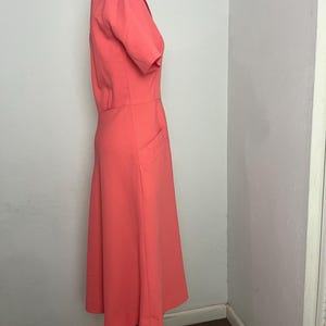 May include: A coral-colored dress with short sleeves and a collared neckline. The dress features a fitted bodice, a defined waist, and a flared skirt. It has two front pockets and is displayed on a mannequin.