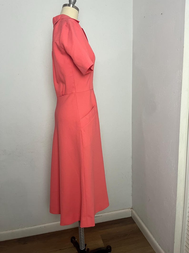 May include: A coral-colored dress with short sleeves and a collared neckline. The dress features a fitted bodice, a defined waist, and a flared skirt. It has two front pockets and is displayed on a mannequin.