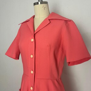 May include: Coral-colored vintage dress with short sleeves, a collared neckline, and a button-down front. The dress features two front pockets and is displayed on a mannequin. The dress is a retro style.