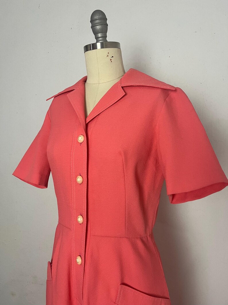 May include: Coral-colored vintage dress with short sleeves, a collared neckline, and a button-down front. The dress features two front pockets and is displayed on a mannequin. The dress is a retro style.