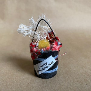 Puede incluir: Un pequeño cubo negro con un forro de tela rojo y blanco y un asa de cuerda blanca. El cubo está lleno de cuerda blanca y una esponja amarilla. El cubo tiene una etiqueta blanca que dice "Odio las tareas domésticas".