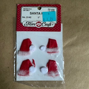 May include: A pack of Fibre Craft Santa hats. The hats are red with white trim and a white pom-pom. The package has a red and white polka dot header with the text "SANTA H" and "1 inch" (2.54 cm).