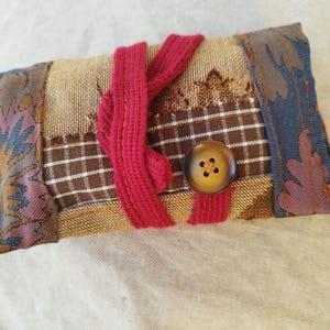 Può includere: Un portafoglio in tessuto fatto a mano con un design patchwork. Il portafoglio presenta un motivo a quadri marrone, motivi floreali e a foglie e una chiusura a nastro rosso. Un grande bottone marrone assicura la chiusura. La combinazione di colori include sfumature di marrone, rosso e blu.