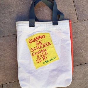 May include: White tote bag with a red side panel and dark blue handles. A yellow patch with red text is sewn onto the front of the bag. The text reads "QUANNO SE SCHERZA BISOGNA ESSE SERI" and "O. DEL GRILLO".