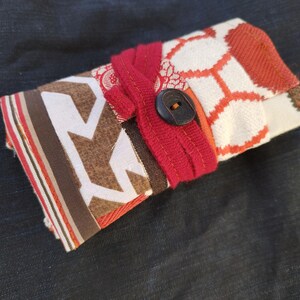 Può includere: Un portafoglio in tessuto fatto a mano con chiusura a bottone. Il portafoglio presenta un design patchwork con vari motivi in rosso, marrone e bianco. Una fascia di tessuto rosso avvolge il portafoglio, fissandolo con un bottone scuro.
