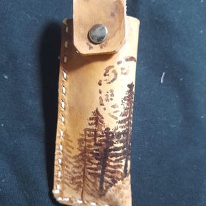 May include: A brown leather case with a flap closure and a metal snap. The case has a design of trees on the front.
