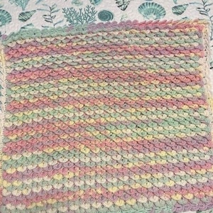 May include: A multi-colored crochet baby blanket with a pastel color scheme of pink, yellow, green, and purple. The blanket is made with a textured stitch that resembles scales.