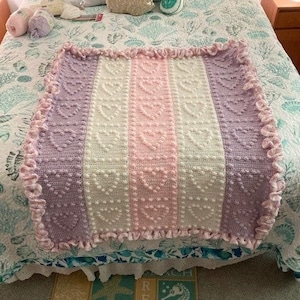 May include: A handmade, pastel-colored baby blanket with a heart pattern. The blanket features vertical stripes of lavender, white, and pink, with a ruffled edge. The blanket is made of soft yarn and is perfect for a nursery or as a gift.