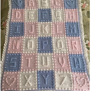 May include: A baby blanket with a crocheted alphabet design in pink, blue, and white. Each letter is surrounded by small, raised, textured circles.