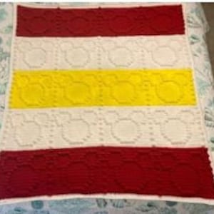 Könnte beinhalten: Eine weiße gehäkelte Babydecke mit roten und gelben Streifen. Die Decke hat ein sich wiederholendes Muster aus kleinen Kreisen.