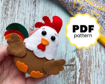Könnte beinhalten: Ein Filz-Hahn-Spielzeug aus braunem, weißem, rotem, orangefarbenem und grünem Filz. Der Hahn hat einen roten Kamm, einen weißen Körper, braune Flügel und orangefarbene Füße. Das Spielzeug wird in einer Hand gehalten. Der Text "PDF pattern" befindet sich in einem weißen Kreis auf gelbem Hintergrund.