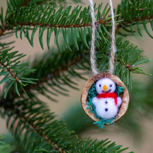 May include: A felt snowman ornament with a red scarf and black eyes, hanging from a branch of a pine tree.